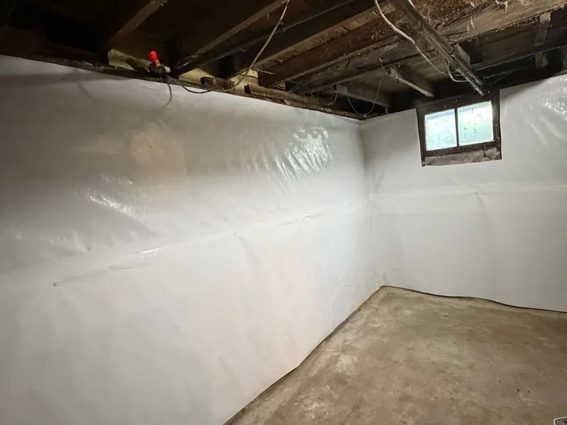 White sheeting covers the walls of a basement, around a small window, over a bare concrete floor.