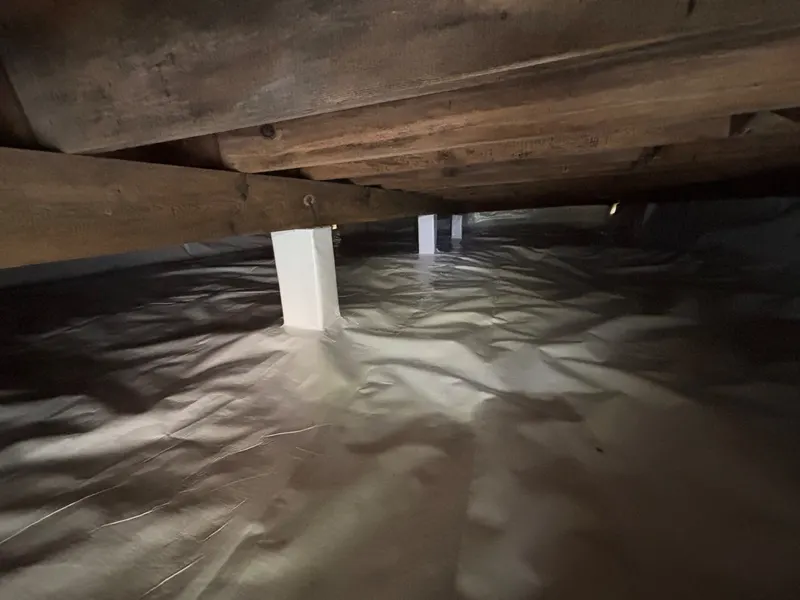 A crawl space with a white liner over the ground and white wrapping around the posts under wood beams.